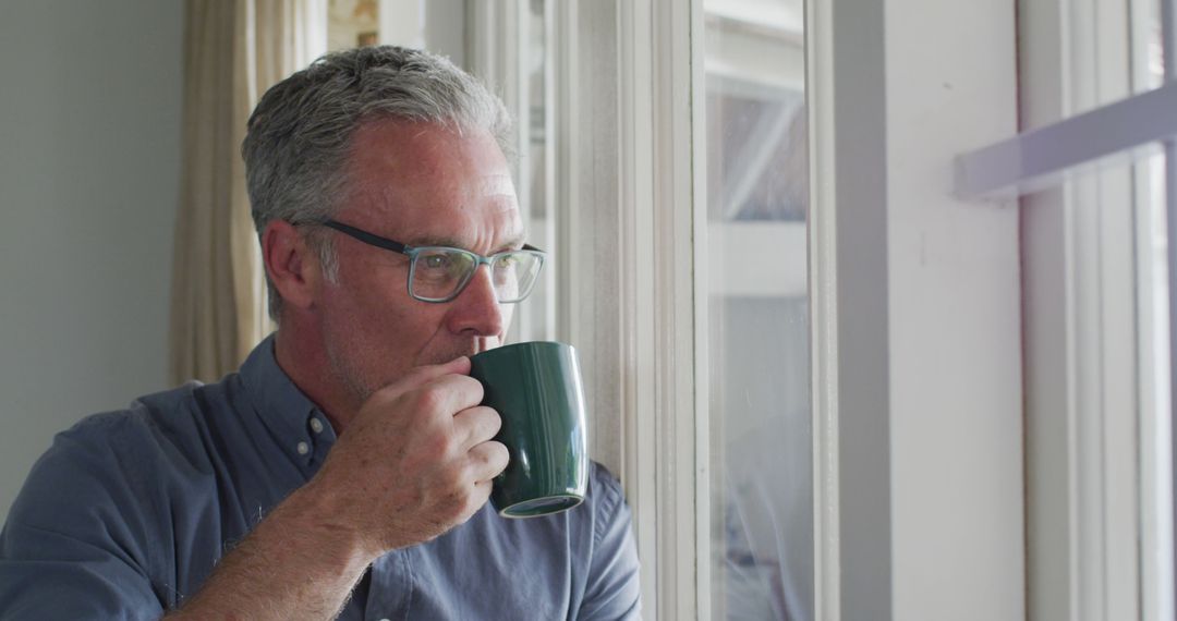 Happy caucasian man wearing glasses, looking through window and drinking coffee - Free Images, Stock Photos and Pictures on Pikwizard.com