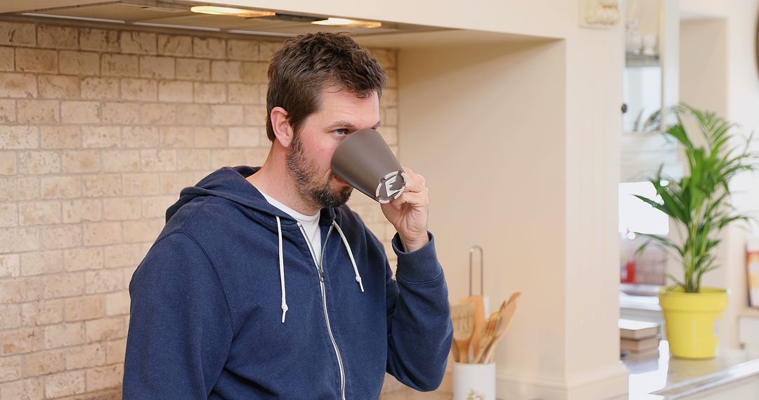 Man drinking coffee in a kitchen wearing a blue hoodie - Free Images, Stock Photos and Pictures on Pikwizard.com