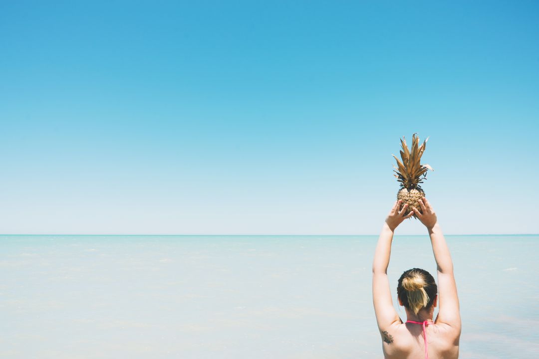 Woman Holding Pineapple Above Head on Sunny Beach - Free Images, Stock Photos and Pictures on Pikwizard.com