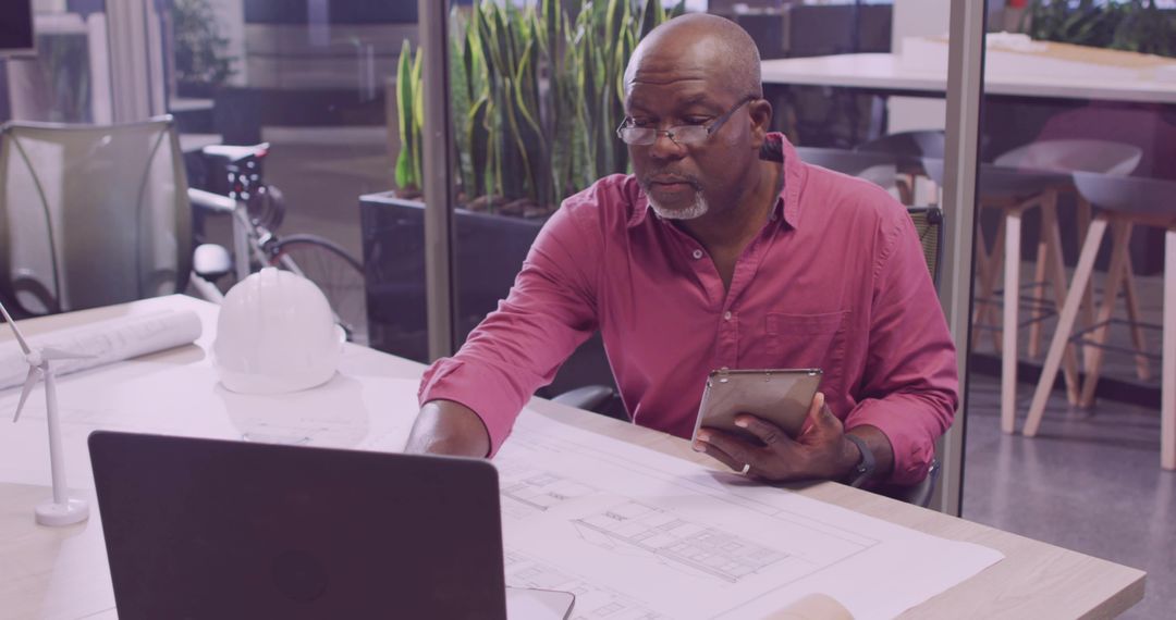 Senior Architect Analyzing Blueprints at Office Desk with Laptop