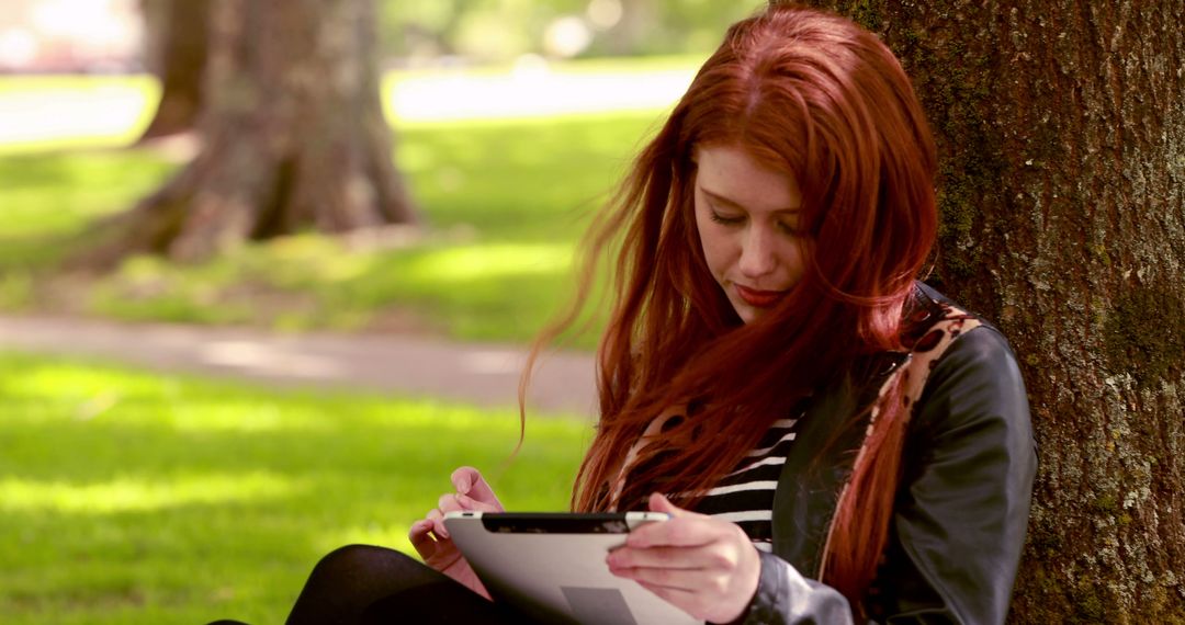 Young woman reading tablet in park under tree - Free Images, Stock Photos and Pictures on Pikwizard.com