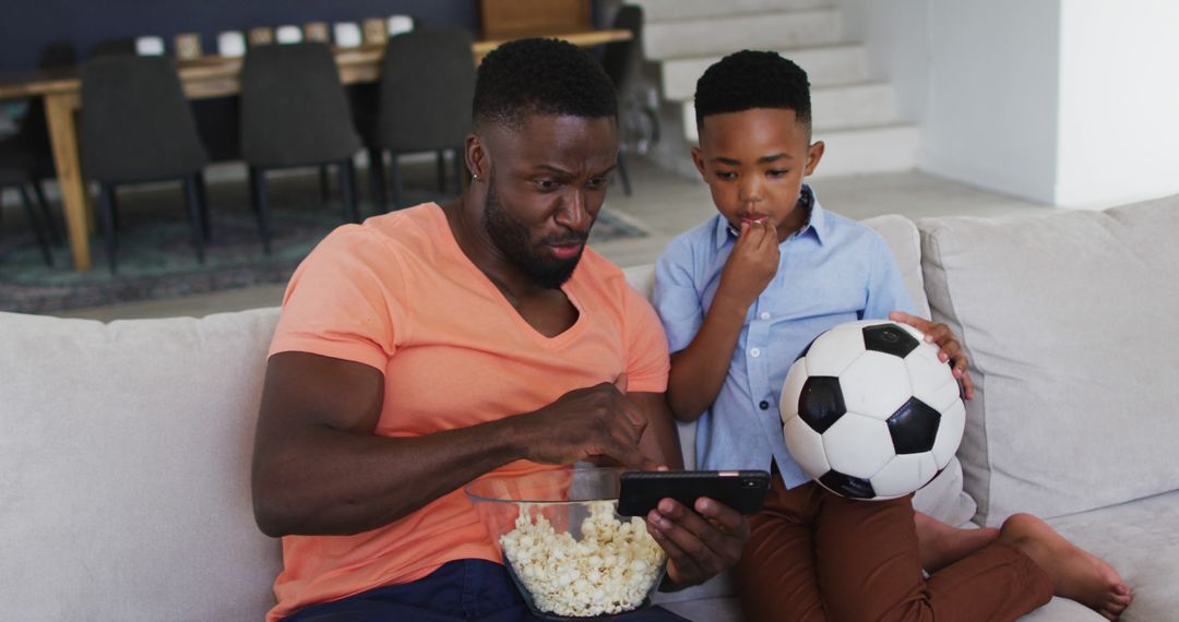 African american father and son eating popcorn and watching a football game on smartphone - Free Images, Stock Photos and Pictures on Pikwizard.com