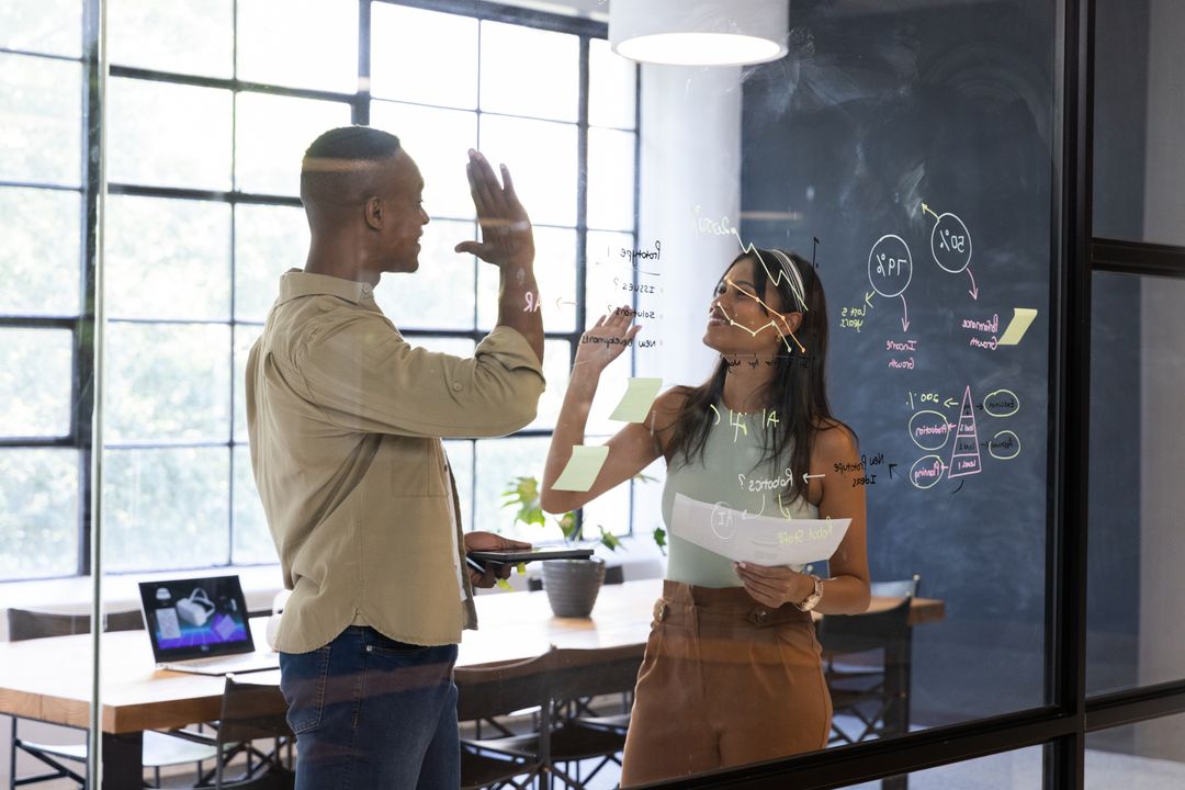 High-fiving while brainstorming on glass wall with sticky notes in modern office meeting