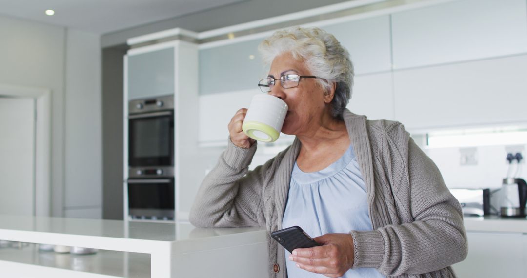 Senior Woman Enjoying Coffee While Using Smartphone in Modern Kitchen - Free Images, Stock Photos and Pictures on Pikwizard.com