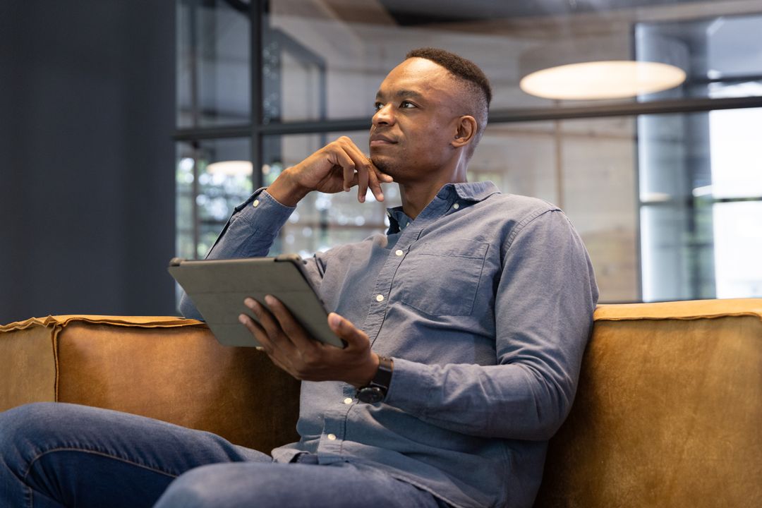 Contemplative African American Professional Using Tablet on Sofa in Office Lounge Casual Business