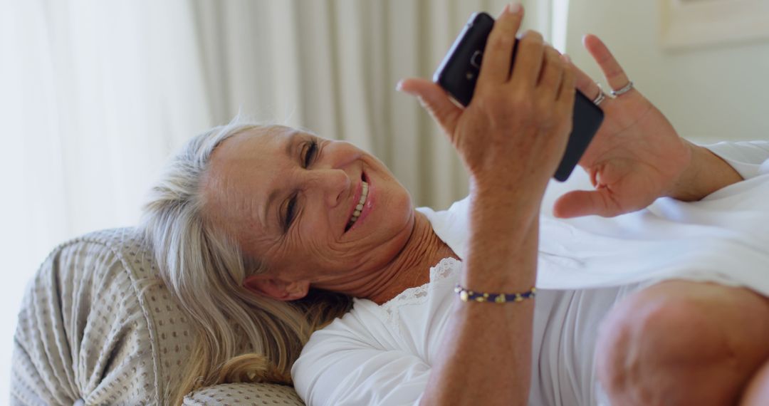 Senior Woman Smiling and Reading on Smartphone While Relaxing in Bed - Free Images, Stock Photos and Pictures on Pikwizard.com