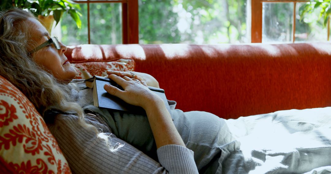 Senior Woman Relaxing on Couch with Book in Sunlit Living Room - Free Images, Stock Photos and Pictures on Pikwizard.com