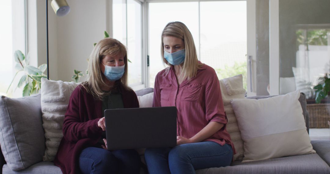 Two Women Wearing Masks Using Laptop on Sofa in Living Room - Free Images, Stock Photos and Pictures on Pikwizard.com