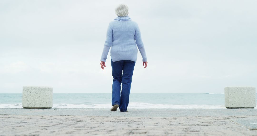 A senior Caucasian woman walks towards the sea on a cloudy day, with copy space - Free Images, Stock Photos and Pictures on Pikwizard.com