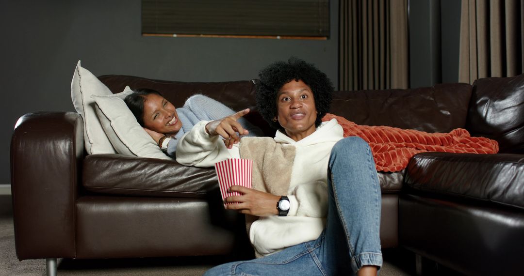 Couple lounging on brown leather sofa sharing popcorn and pointing at screen