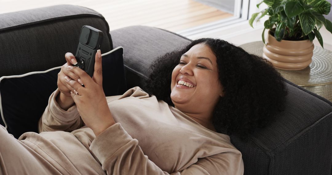 Laughing woman reclining on sofa holding smartphone in cozy loungewear relaxing at home