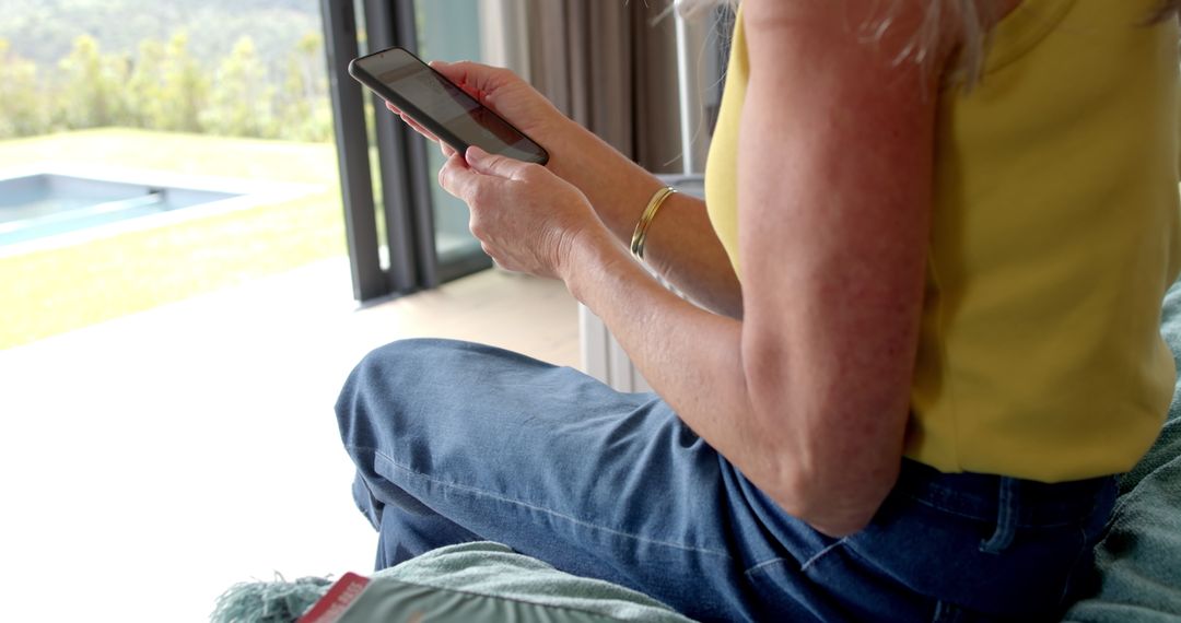 Mature Woman Sitting by Window Using Smartphone in Relaxed Setting