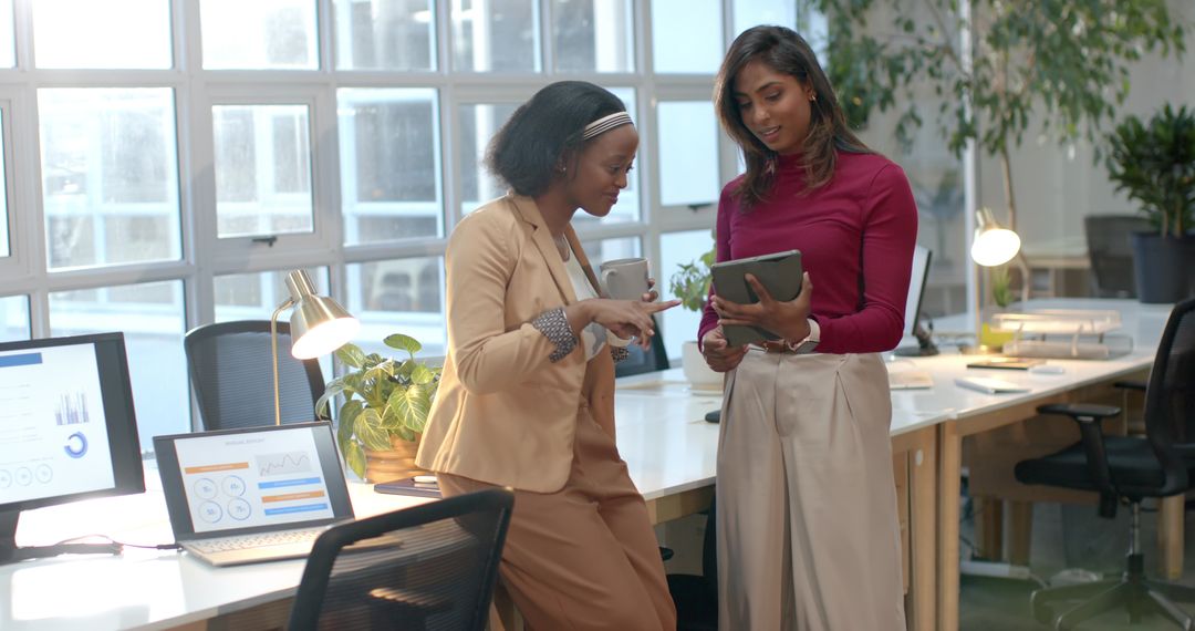 Two diverse female coworkers collaborating and discussing project on tablet in sunlit office