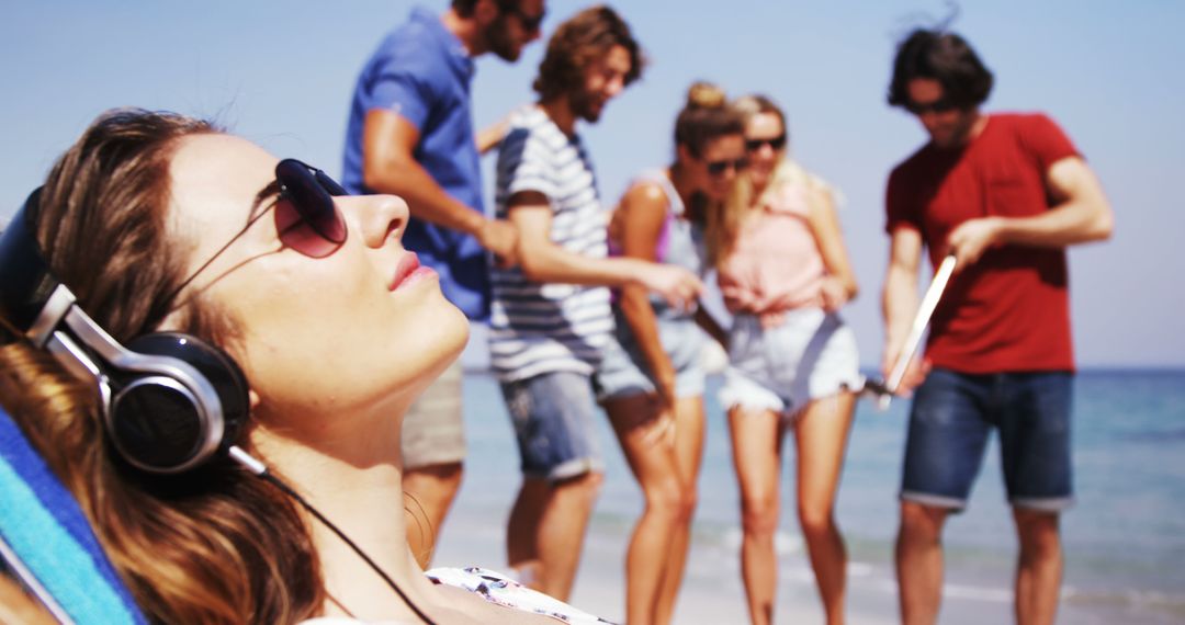 Relaxing Woman with Headphones Lounging by Ocean, Group of Friends in Background - Free Images, Stock Photos and Pictures on Pikwizard.com