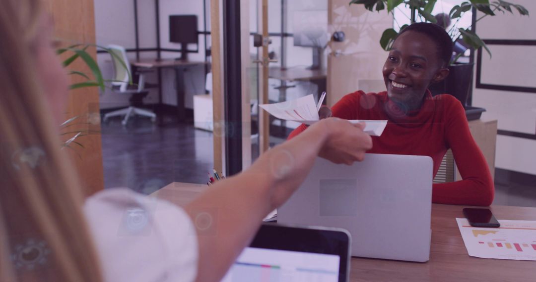 Smiling colleague handing printed report across desk with laptop in modern office