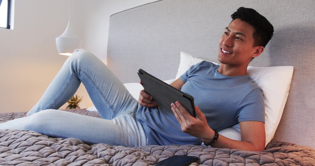 Smiling Young Asian Man Relaxing on Bed with Tablet at Home
