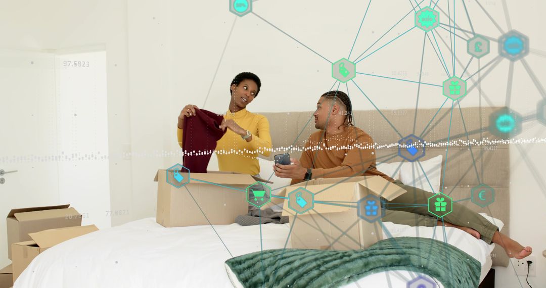 Couple unpacking boxes in cozy bedroom holding sweater and using smartphone