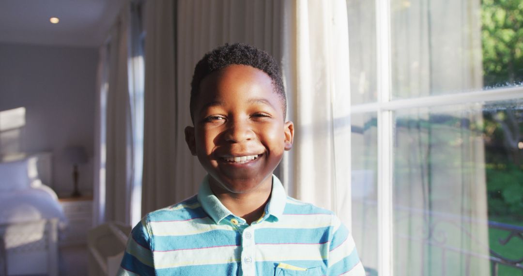 Smiling African American Boy in Striped Shirt Near Sunlit Window - Free Images, Stock Photos and Pictures on Pikwizard.com