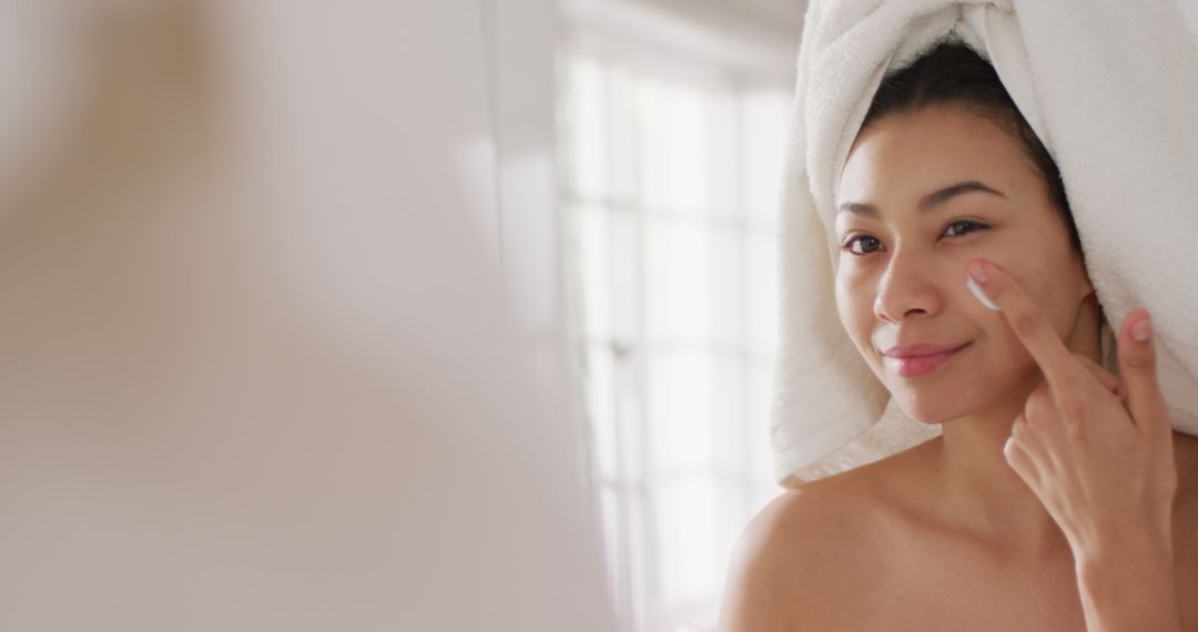 Image of portrait of smiling biracial woman with towel on hair applying cream in bathroom - Free Images, Stock Photos and Pictures on Pikwizard.com