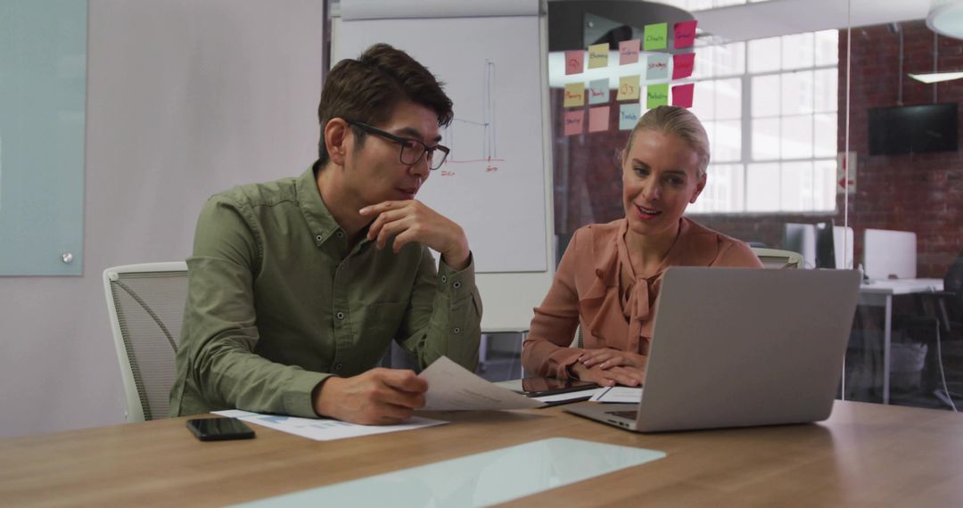 Two Colleagues Discussing Project Over Laptop and Documents in Modern Glass Office Meeting