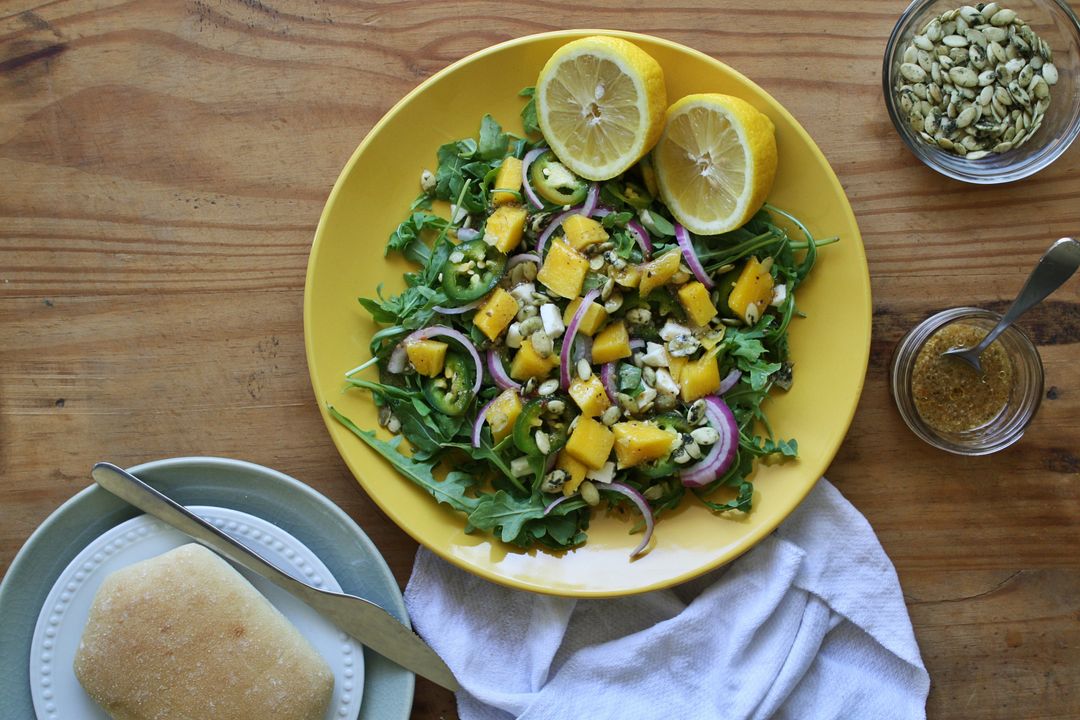 Serving vibrant arugula mango salad with lemon halves, red onion and pumpkin seeds
