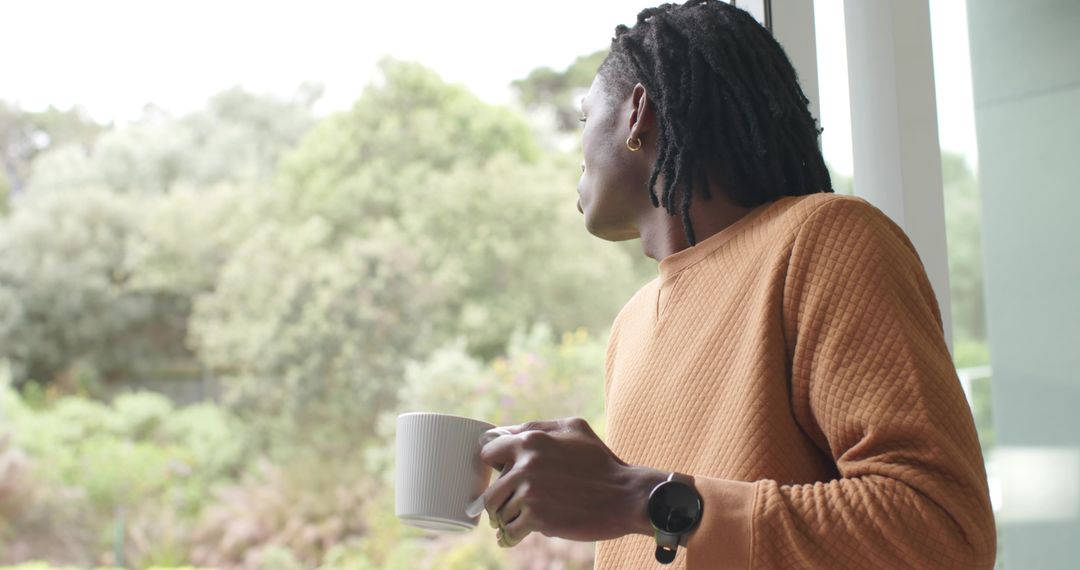 African American man standing by glass window holding mug in rust sweater morning calm