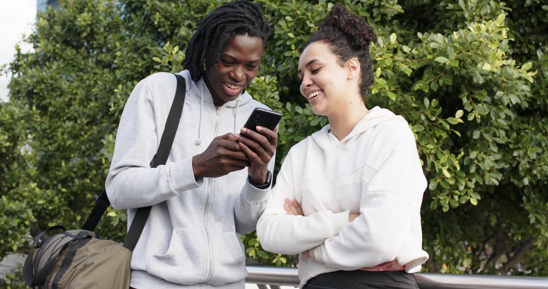 Multicultural students checking smartphone on campus railing, smiling in casual hoodies