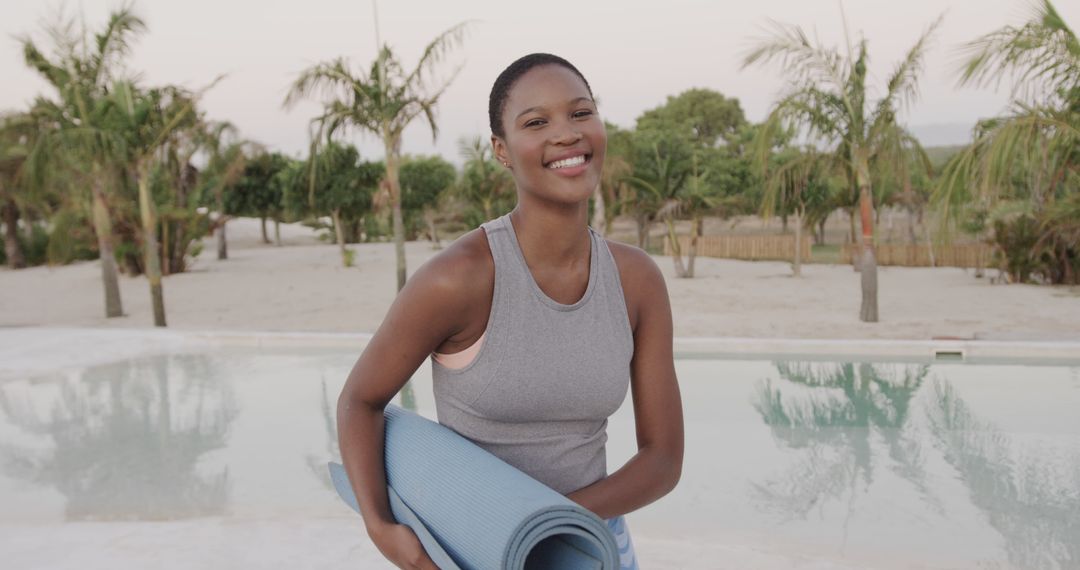 Smiling woman holding yoga mat near pool with palm trees in background - Free Images, Stock Photos and Pictures on Pikwizard.com