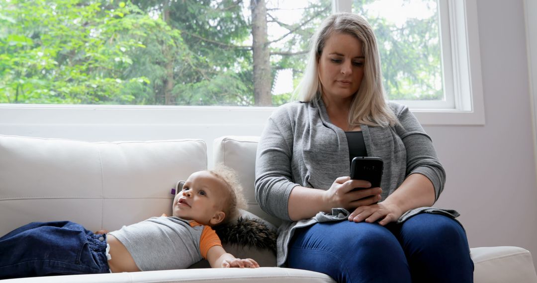 Mother Using Smartphone While Sitting with Toddler on Couch in Bright Living Room - Free Images, Stock Photos and Pictures on Pikwizard.com