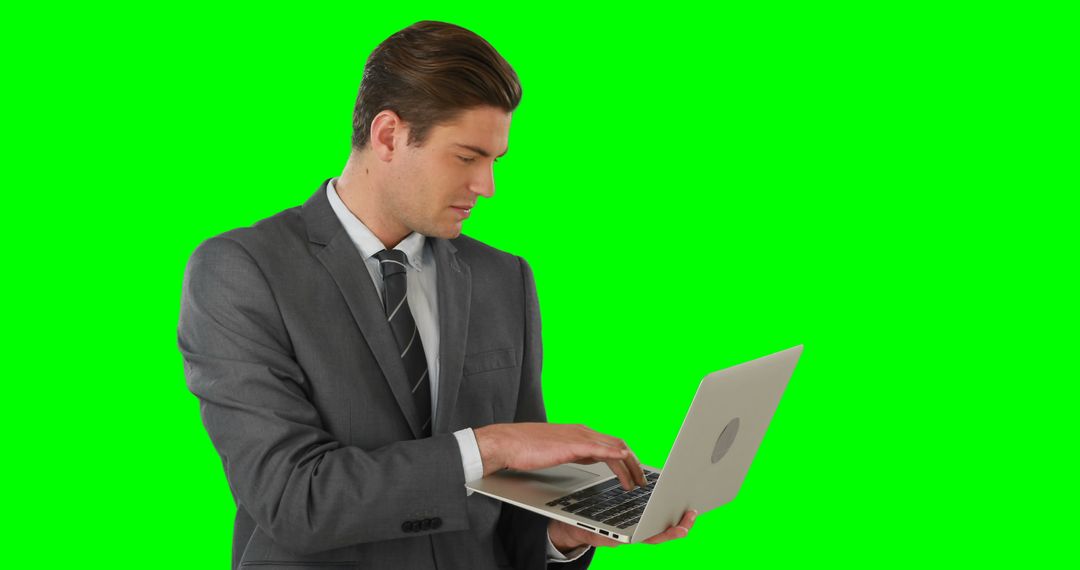 A focused young businessman in a gray suit works intently on his laptop against a green backdrop. - Free Images, Stock Photos and Pictures on Pikwizard.com