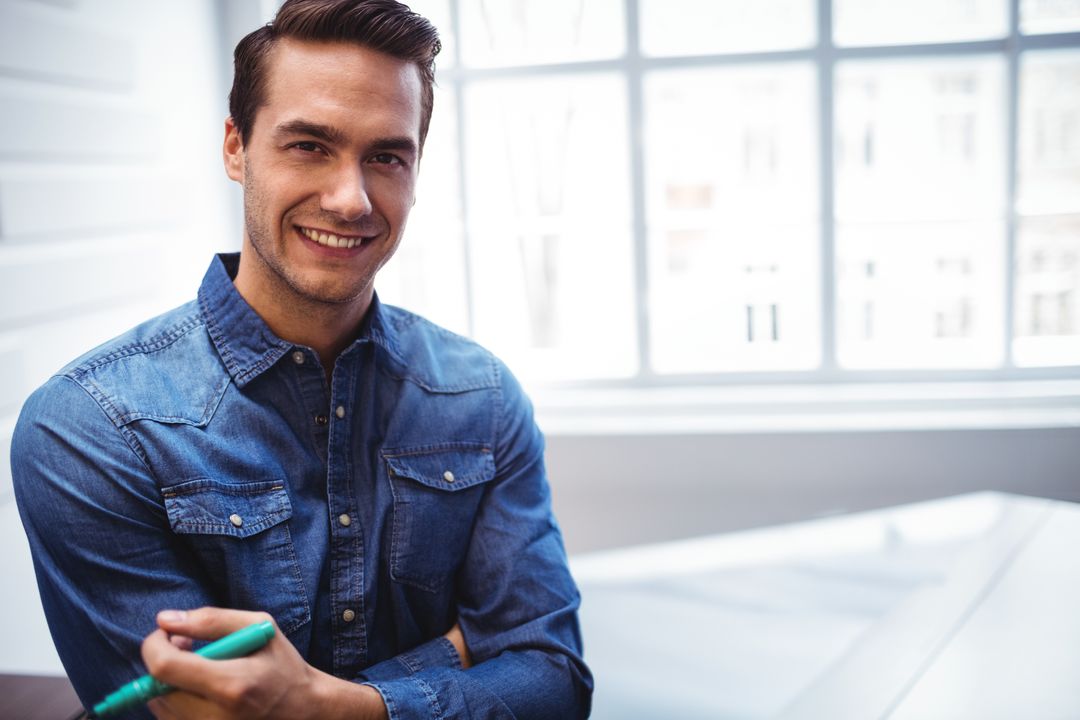 Smiling Young Man in Denim Holding Modern Device in Office Space
