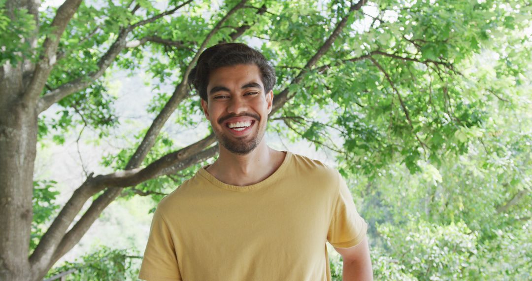 Portrait of cheerful biracial man in yellow t-shirt is standing against trees - Free Images, Stock Photos and Pictures on Pikwizard.com