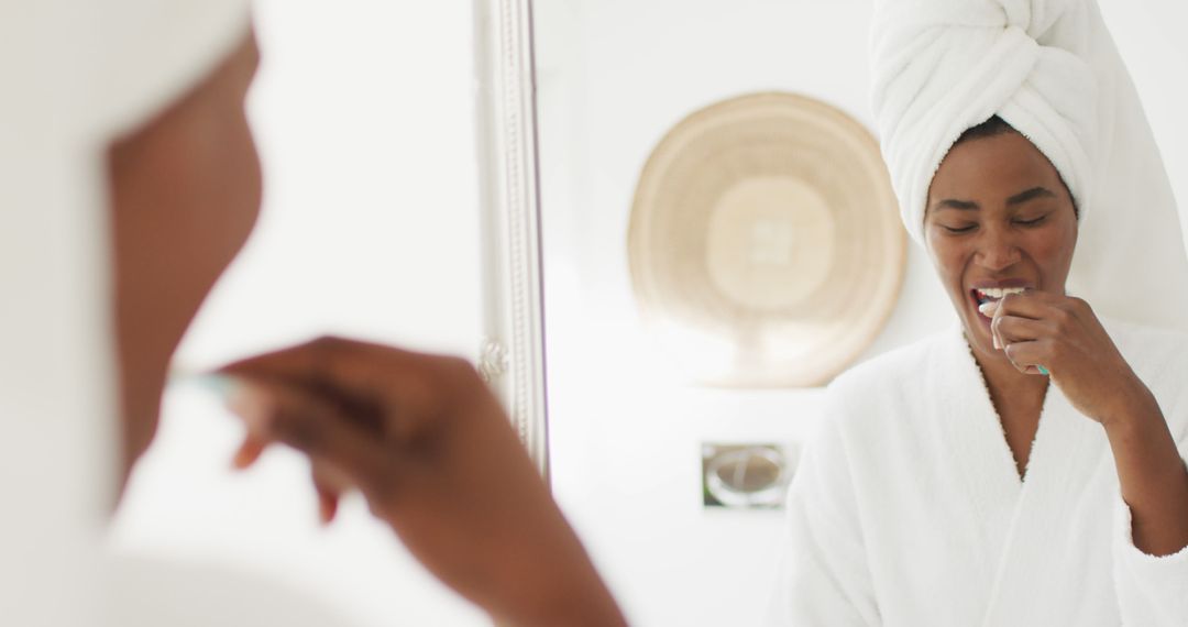 Image of happy african american woman in robe looking at mirror and brushing teeth - Free Images, Stock Photos and Pictures on Pikwizard.com