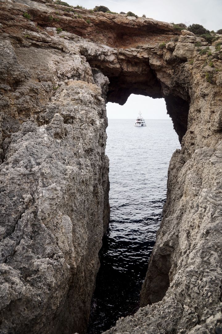 Yacht visible through rocky arch on Mediterranean coastline - Free Images, Stock Photos and Pictures on Pikwizard.com