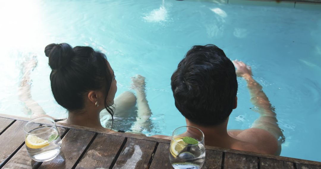 Diverse couple relaxing at pool edge with lemon-mint drinks on wooden deck
