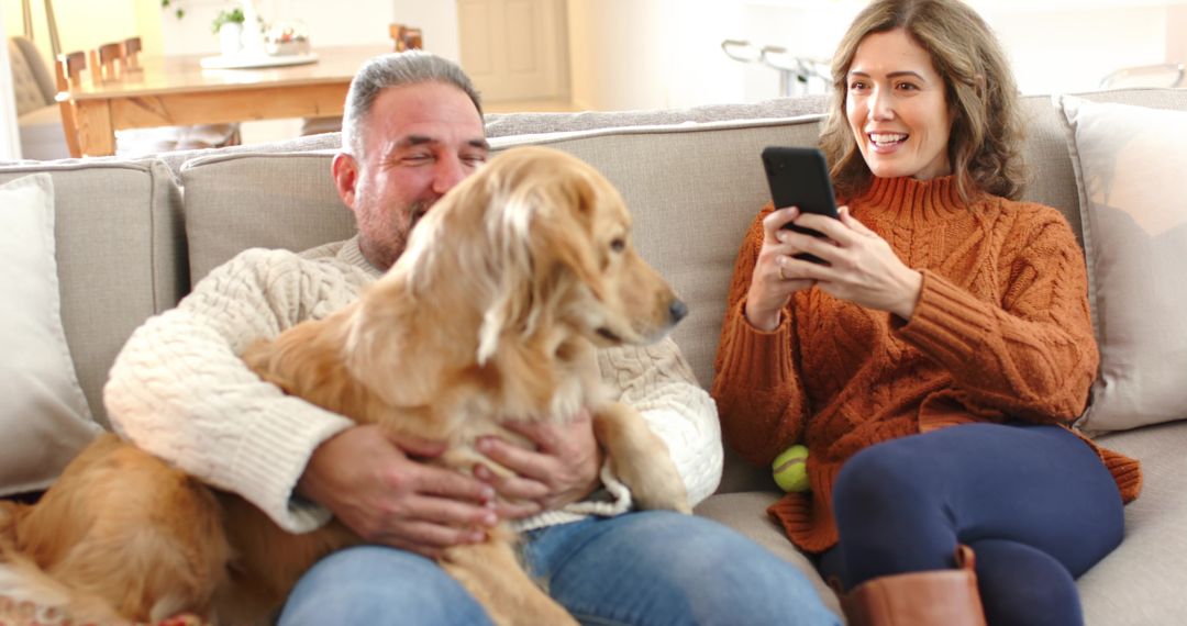Couple relaxing on sofa with golden retriever while woman using smartphone and smiling