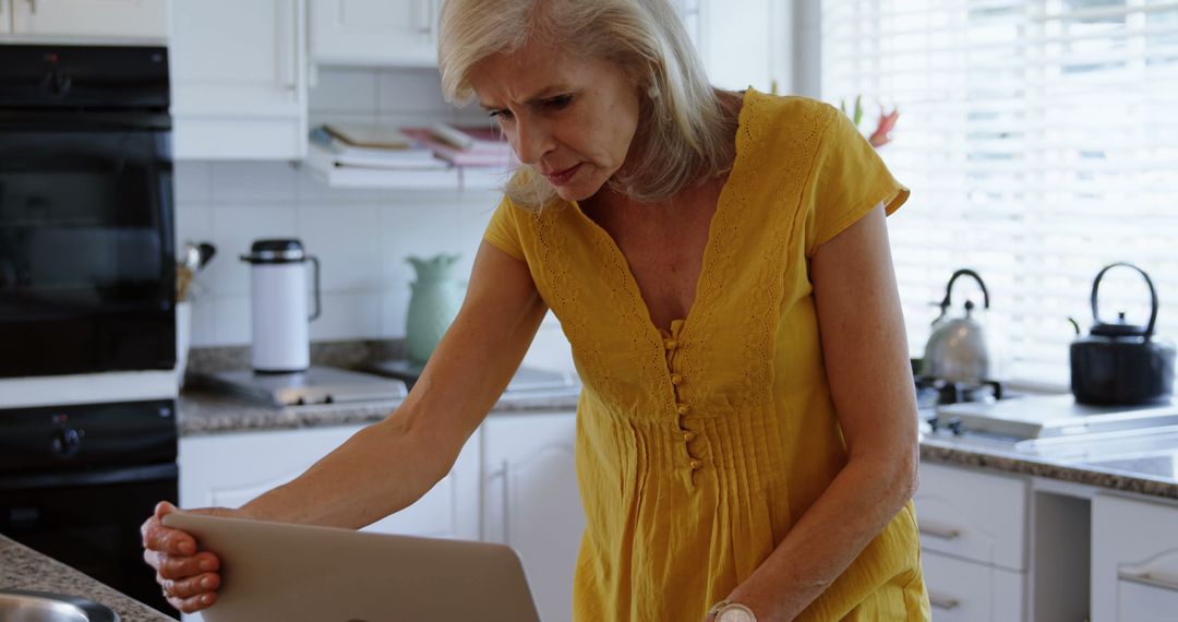 Senior Woman Using Laptop in Kitchen Wearing Yellow Dress - Free Images, Stock Photos and Pictures on Pikwizard.com