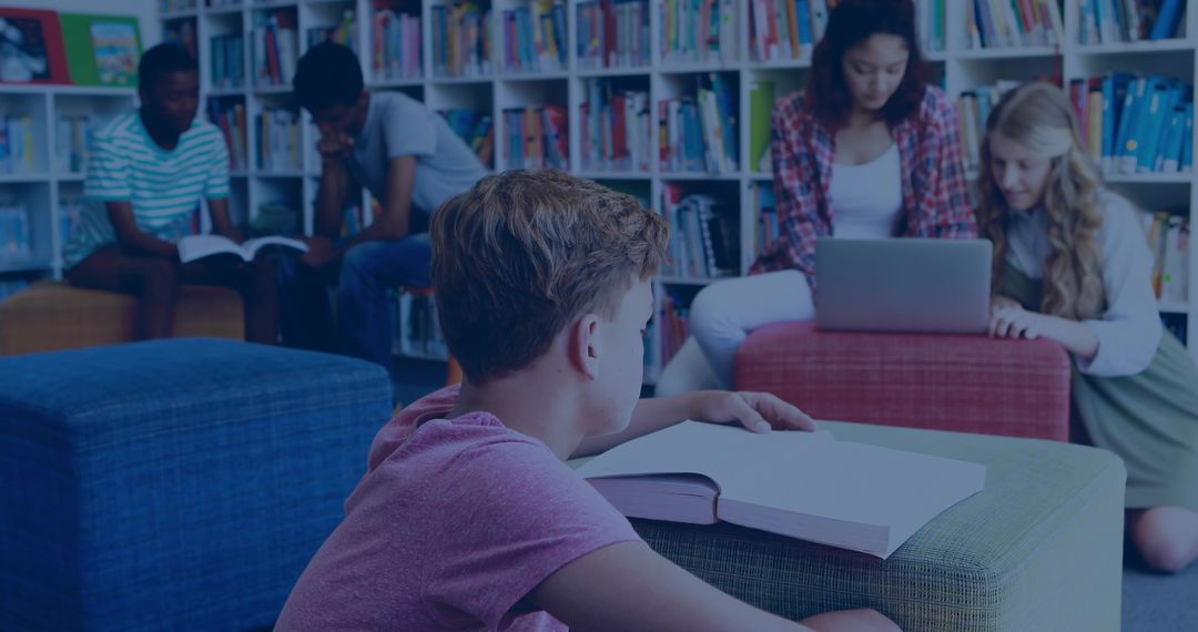 Teen reading textbook on library ottoman while classmates studying with laptop and books
