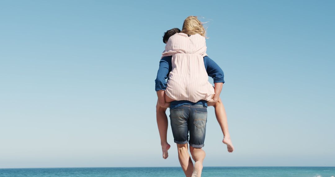 Couple Enjoying Piggyback Ride by Sea Shore Under Clear Blue Sky - Free Images, Stock Photos and Pictures on Pikwizard.com