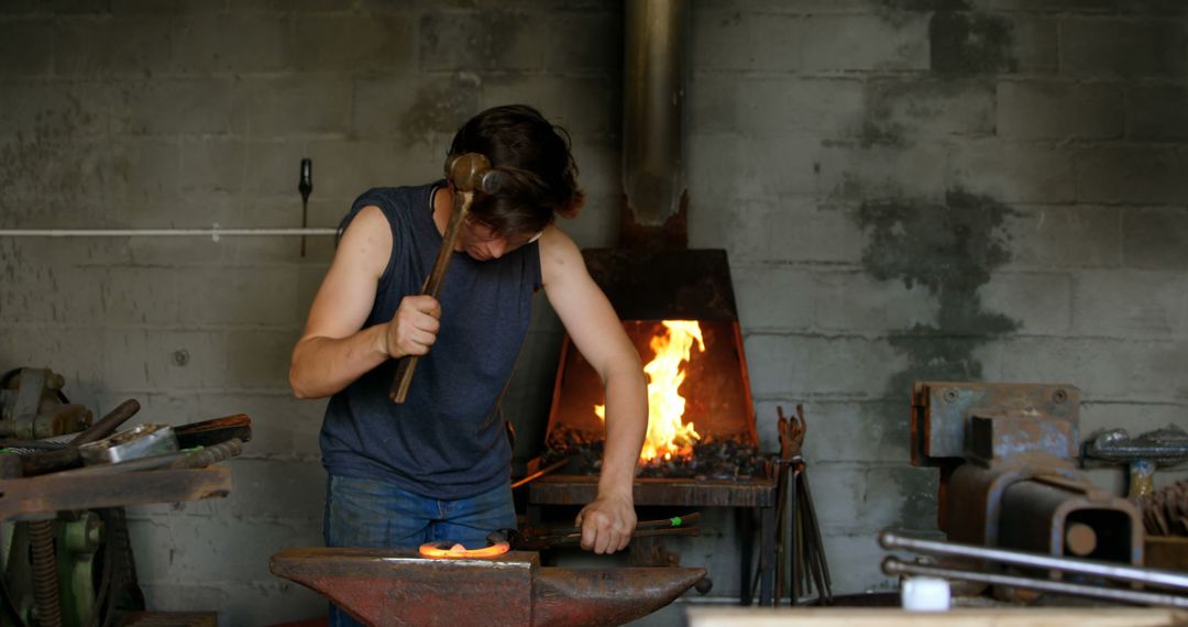 Blacksmith Wearing Sleeveless Shirt Hammering Glowing Metal in Workshop - Free Images, Stock Photos and Pictures on Pikwizard.com