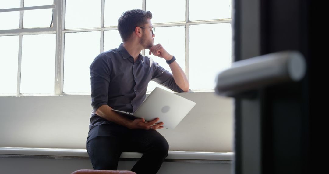 Contemplative Young Professional with Laptop by Office Window