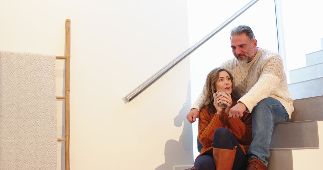 Couple sitting on stairs holding mug wearing cozy knit sweaters with warm window light