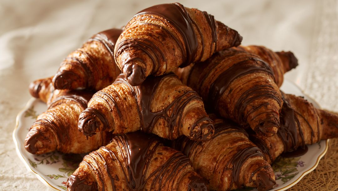 Stacked chocolate-drizzled croissants on vintage floral plate, golden buttery flaky layers