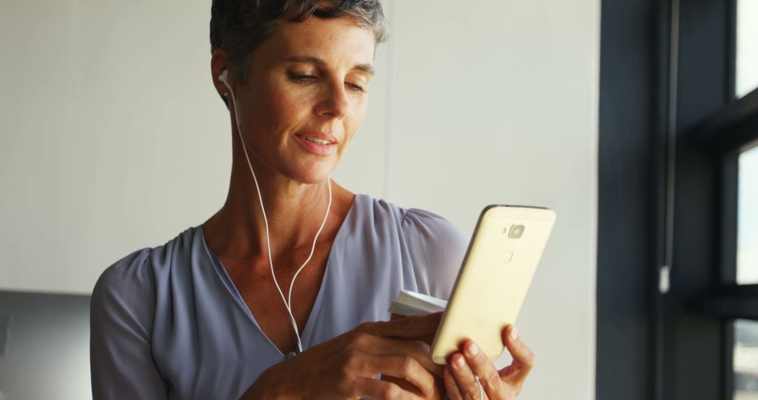 Older Woman Connecting Online Using Smartphone and Earphones in Modern Office - Free Images, Stock Photos and Pictures on Pikwizard.com