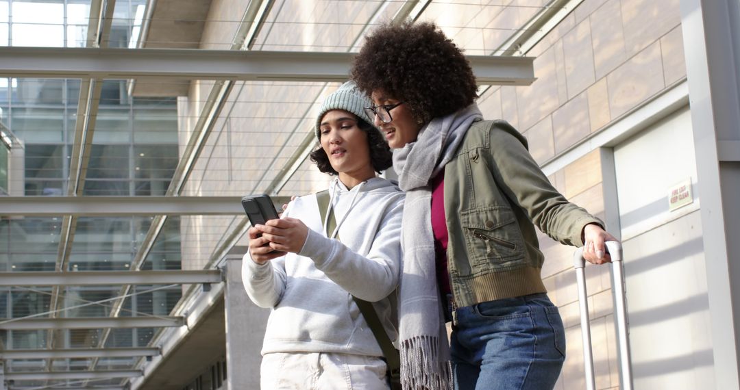 Two friends checking smartphone while holding rolling suitcase at modern transit terminal