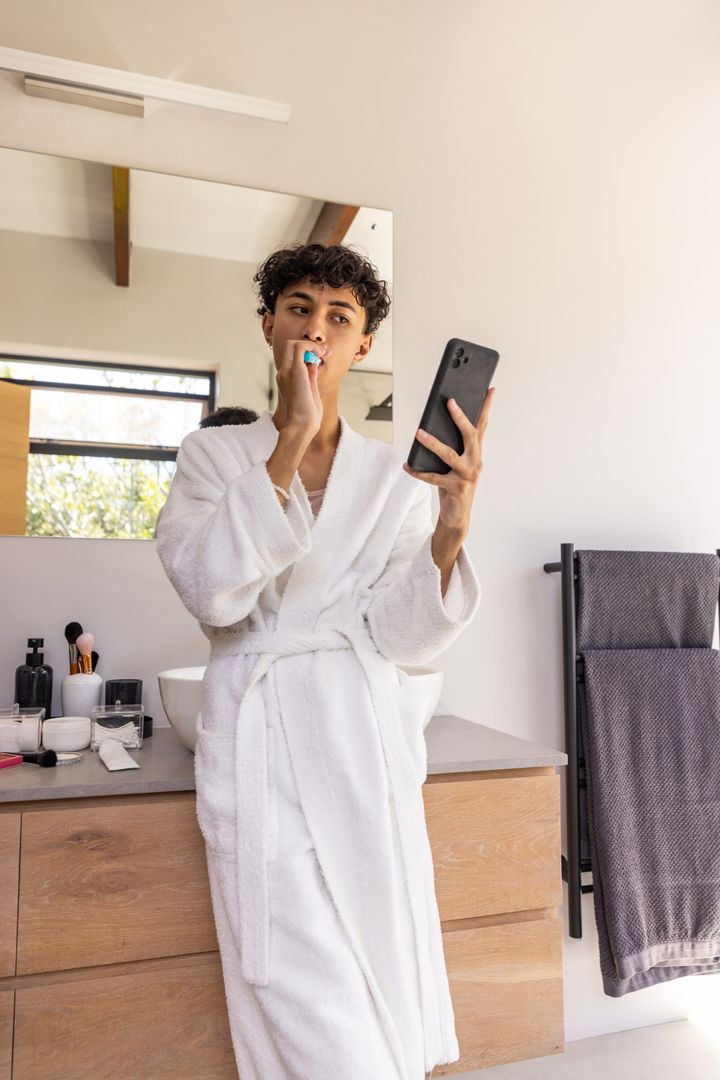Young Man in Bathrobe Brushing Teeth and Using Smartphone