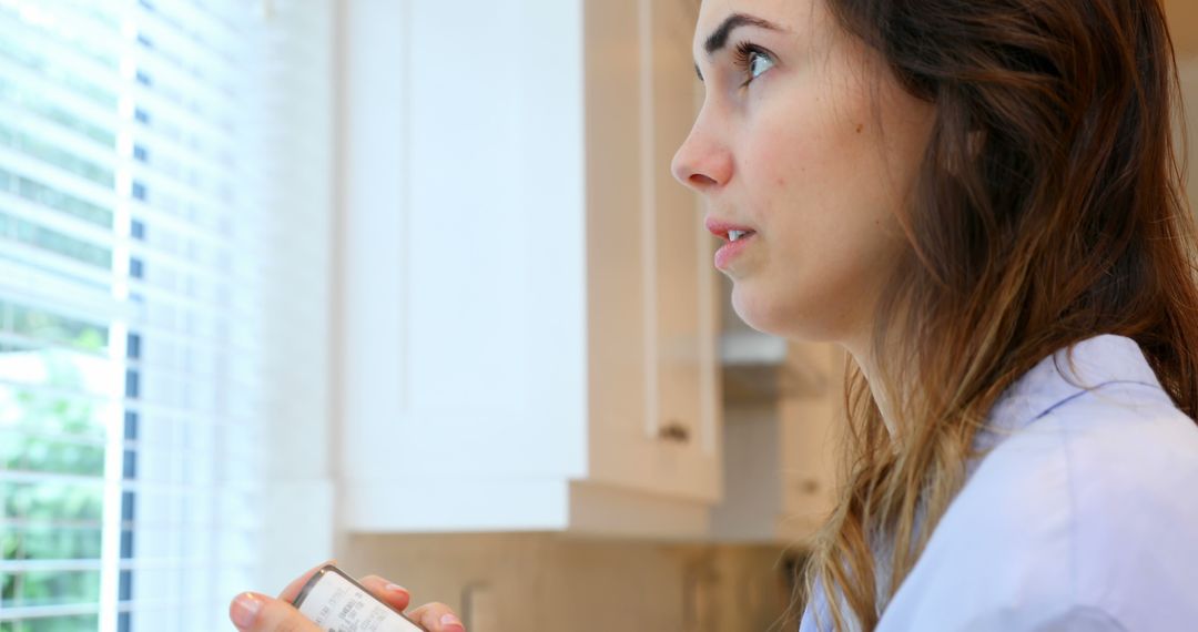 Woman contemplating while looking outside holding smartphone in kitchen - Free Images, Stock Photos and Pictures on Pikwizard.com