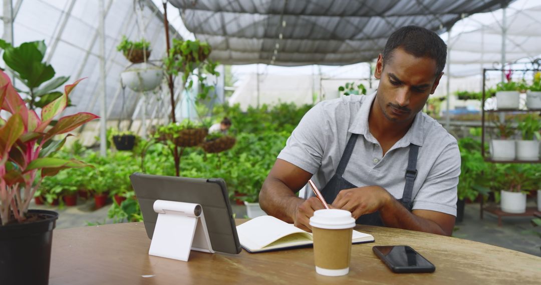 Focused Botanist Writing Notes in Greenhouse with Tablet and Coffee