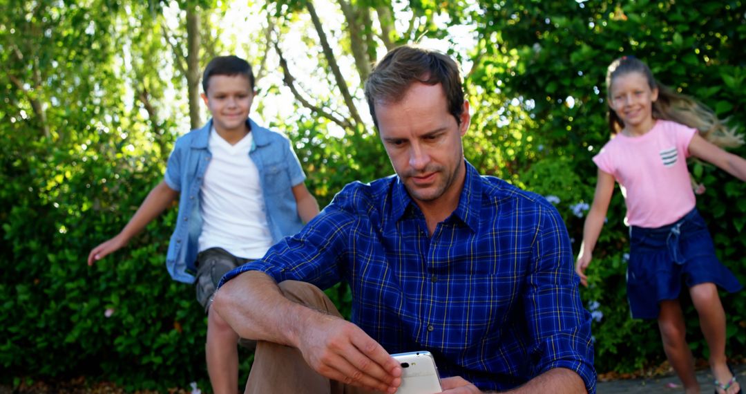 Father Takes a Break with Phone While Kids Play in Garden - Free Images, Stock Photos and Pictures on Pikwizard.com