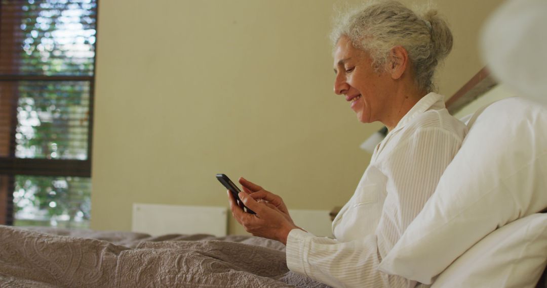 Senior woman smiling while using smartphone in bed on sunny morning - Free Images, Stock Photos and Pictures on Pikwizard.com